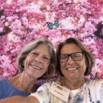 Two smiling older women pose for a selfie in front of a vibrant pink floral background with butterflies. One wears glasses and a name tag.