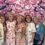 Six women stand together smiling in front of a vibrant floral backdrop with pink blossoms and butterflies.