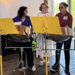 Three women stand behind steel drums with yellow music stands, smiling and talking indoors near a sunlit window.
