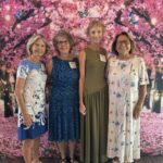 Four women stand side by side, smiling, in front of a backdrop featuring pink cherry blossoms, butterflies, and string lights. They are wearing name badges and summer dresses.