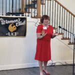 A woman in a red dress speaks at a microphone in front of a staircase and a banner that reads "The Women's Club at UCF.