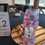 A round mirror with a floral-decorated glass, a Table 12 sign, and empty champagne flutes on a black tablecloth in a bright room.