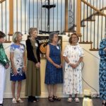 Six women stand in a line indoors, five facing forward and one reading from a paper near a microphone. A banner and staircase are visible in the background.