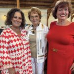 Three women stand indoors, smiling at the camera; one wears a red and white patterned dress, one a white jacket, and one a solid red dress.