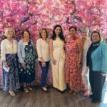 Seven women stand in front of a vibrant pink floral backdrop with butterflies, smiling at the camera. They are dressed in colorful, semi-formal attire.