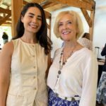 Two women stand side by side indoors, both smiling at the camera. The younger woman wears a sleeveless beige top, and the older woman wears a white blouse and patterned blue skirt.