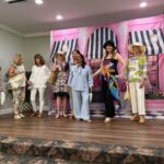 Seven women stand on a stage in front of a pink backdrop with black and white stripes, wearing a variety of summer outfits and hats, while another woman stands to the far right.