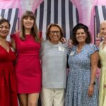 Five women stand together smiling in front of a pink backdrop with black and white striped awnings and columns, wearing brightly colored dresses.