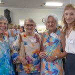 Four women wearing name tags pose together at a daytime indoor social event with other attendees in the background.