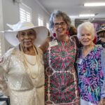 Three older women, dressed in colorful, patterned outfits and name badges, stand together smiling at an indoor social event.