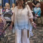 A woman in pastel clothing walks and smiles at a seated group of older adults during an indoor social event.