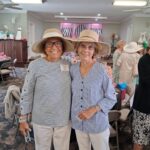 Two older women in straw hats and striped shirts stand together, smiling, at a social gathering in a decorated room with chairs and people in the background.