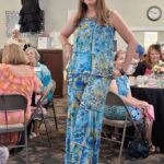 A woman in a blue patterned outfit poses and smiles at an indoor event, with seated people and tables decorated with flowers in the background.