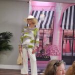 A woman wearing a wide-brim hat, patterned top, white pants, and sandals stands on a stage with a boutique backdrop.