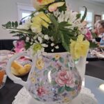 A floral teapot vase filled with assorted flowers sits on a lace doily, with people and tableware visible in the background.