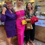 Three women stand together in a shop, smiling. One wears a purple outfit, one a bright pink dress with a yellow purse, and the third wears a black outfit with sandals. Jewelry displays are visible behind them.