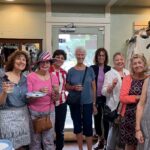 Eight women stand together inside a boutique, smiling at the camera, several holding drinks, with clothing and accessories displayed around them.