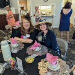 Two women in hats sit at a decorated table set for tea, smiling at the camera. Other people are standing and talking in the background. The table has pink napkins and lace placemats.