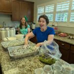 Two women in a kitchen prepare food; one mixes a tray of salad by hand, while the counter holds pitchers of milk, grapes, desserts, and stacked dishware.