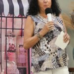 Woman with curly dark hair holds a microphone and a notecard, standing in front of a backdrop featuring a striped awning, flowers, and a window display.
