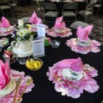 Round table with floral placemats, teacups, pink napkins, gold utensils, a teapot with flowers, lemon wedges, sugar packets, and menu cards; folding chairs surround the table.