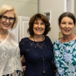 Three women stand together indoors, smiling at the camera; one wears glasses and a white top, the others wear dark blue and blue floral tops.