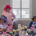 Three women wearing colorful hats sit and stand around a table set for tea, with floral decorations, teacups, and name tags, in a bright room with large shuttered windows.